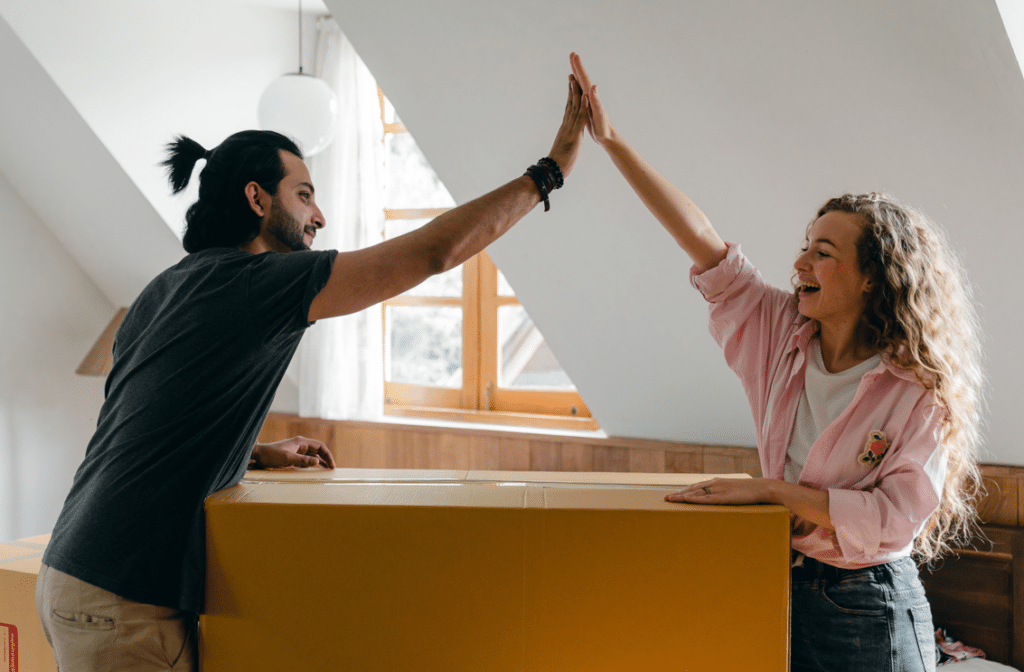 Um homem e uma mulher comemorando em um apartamento. Eles estão sorrindo e segurando uma caixa de mudança.
