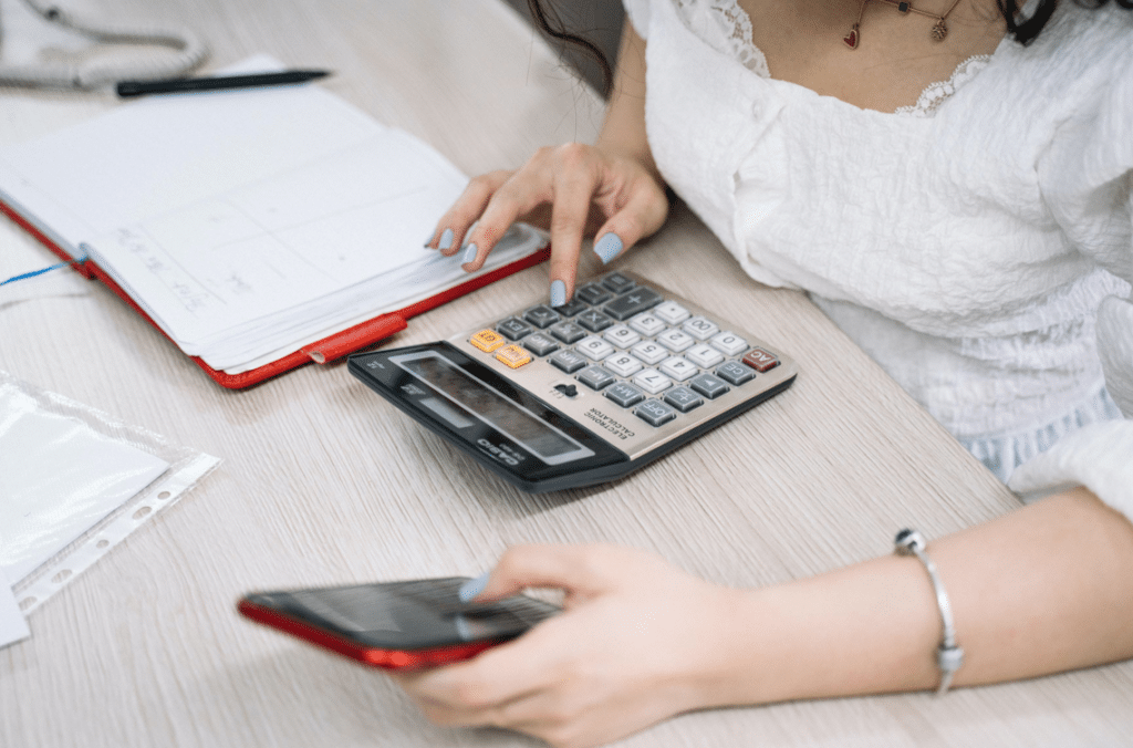 Mulher usando uma calculadora sobre uma mesa, enquanto segura um celular em outra mão. Sobre a mesa, há um caderno vermelho aberto, uma caneta e folhas.