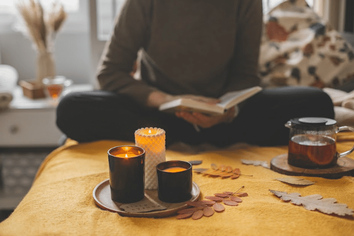 Pessoa sentada na sala de estar lendo um livro. Na mesa tem velas e chá.