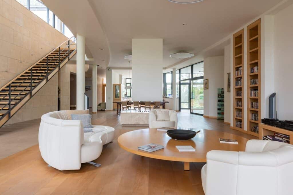 Sala de estar ampla, com piso de madeira, mesa de centro circular do mesmo material e sofás e poltronas brancos. Decoração sustentável também é sinônimo de exclusividade